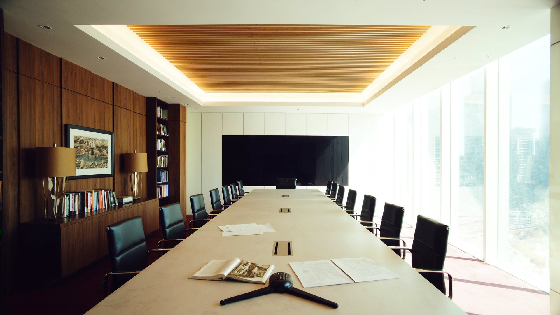 The People in the Loop boardroom in daylight, seen from the doorway: a long table running away from you, an open book and a conference phone on it, walnut shelving on the left, a screen on the far wall, daylight through the window wall.
