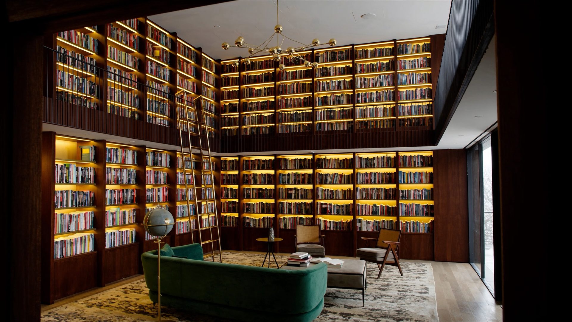 A two-storey library inside an office, seen from the doorway: shelves of books lit warm from within run up both walls past a mezzanine, a brass ladder leans against them, and a green velvet sofa, armchairs and a low table sit on a rug in front.